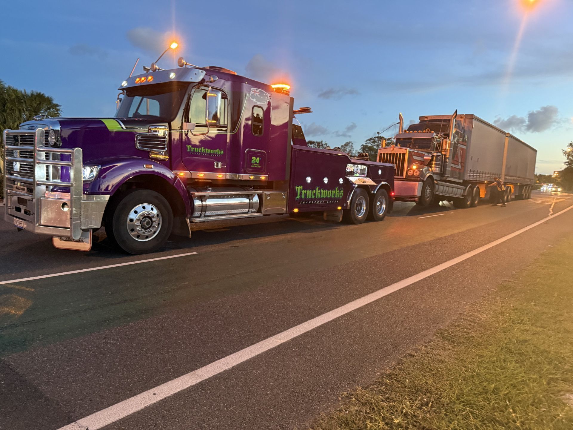 Purple tow truck towing a semi-truck with a trailer on a road at dusk. Truck has lights on.— Fitzroy Towing & Transport in Parkhurst, QLD