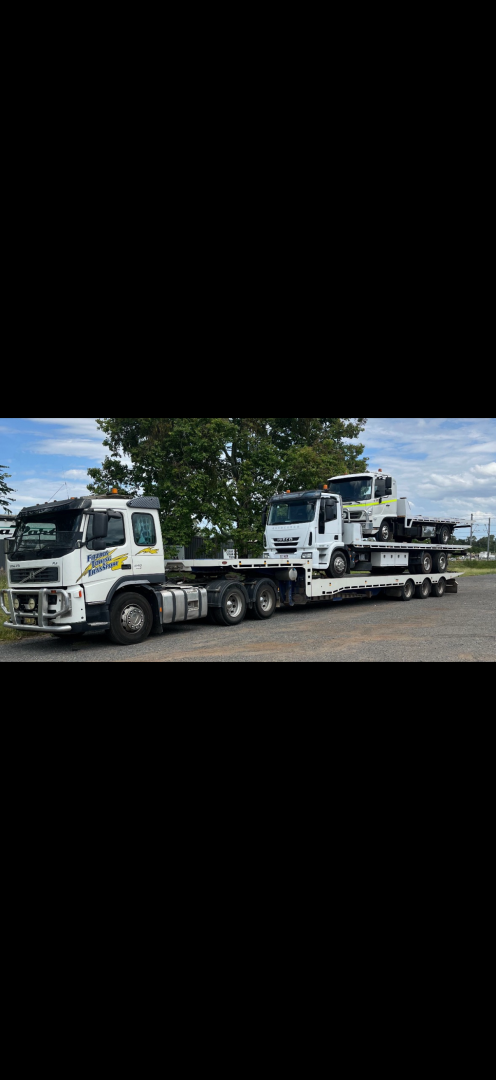 Truck hauling two other trucks on a flatbed trailer, parked on asphalt near trees.— Fitzroy Towing & Transport in Parkhurst, QLD