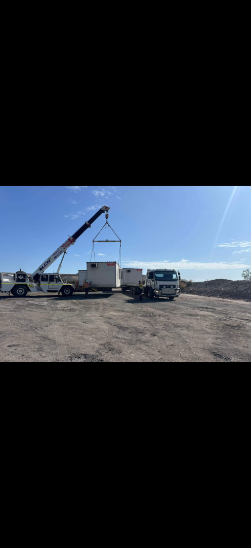 A crane lifting a white structure between two trucks on a dirt lot under a blue sky.— Fitzroy Towing & Transport in Parkhurst, QLD