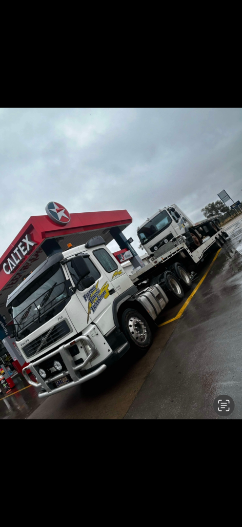 A white tow truck with a smaller vehicle on its flatbed is parked near a Caltex gas station on a wet day.