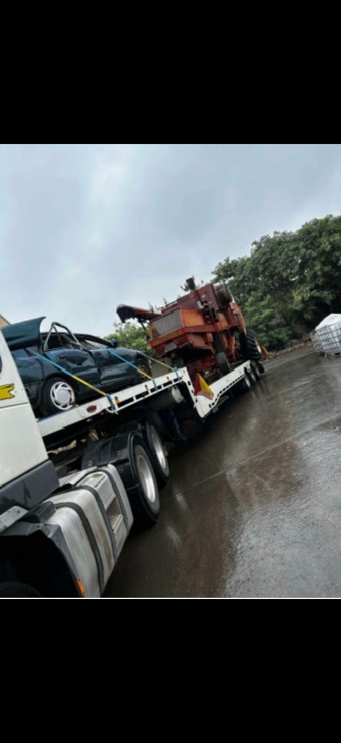 Tow truck carrying a car and a large, rusty piece of machinery on a wet road.
