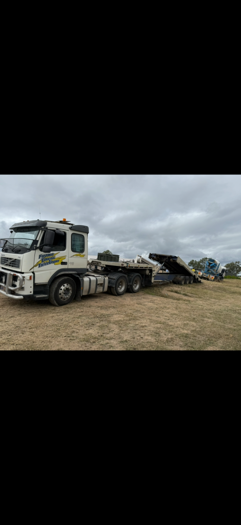 White semi-truck with a flatbed trailer on a dirt field under a cloudy sky.