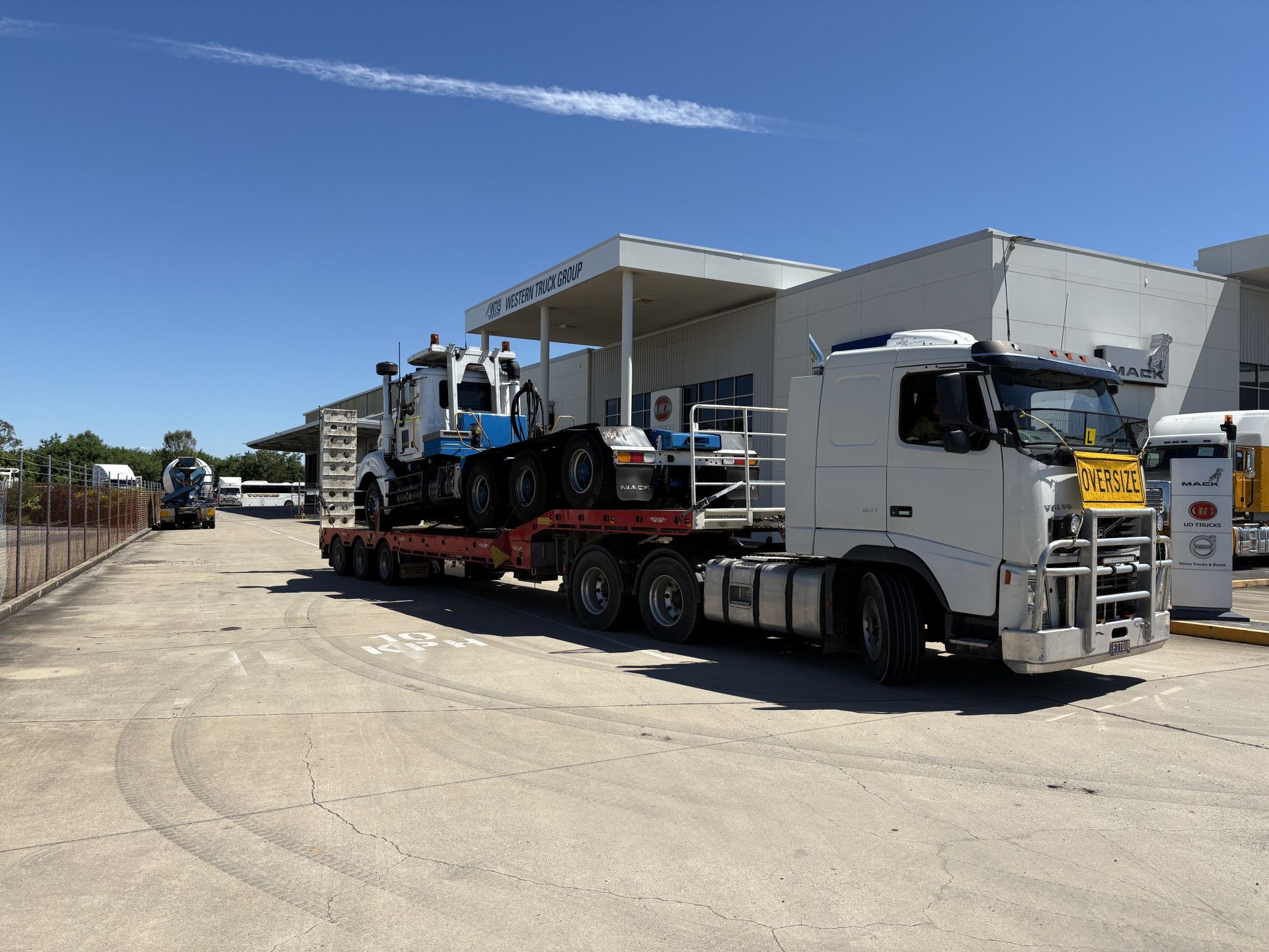Truck transporting a blue and white machine on a flatbed trailer near a building on a sunny day.— Fitzroy Towing & Transport in Parkhurst, QLD