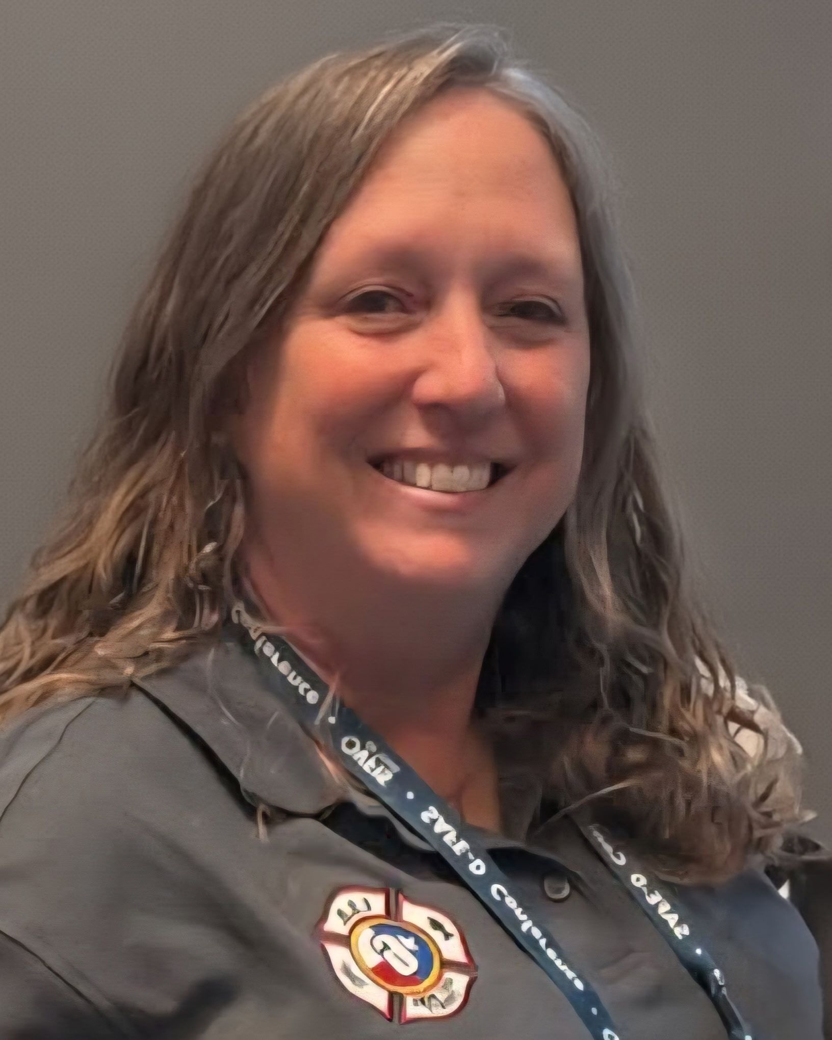 Woman with long wavy hair smiles, wearing a gray shirt with a badge, possibly at a conference.