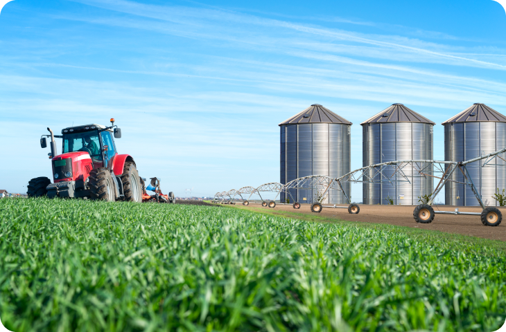 Um trator está arando um campo com silos ao fundo.