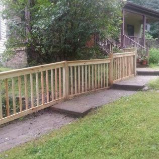 Wooden fence along a concrete path leading to a house with stairs; lush green grass and trees surround.