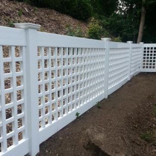 White lattice fence along a dirt area, separating a yard from a hill with greenery.