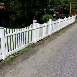 White picket fence lining a road, with green grass and trees in the background.