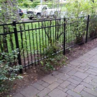 Black metal fence bordering a sidewalk with greenery behind it, a truck in the background.