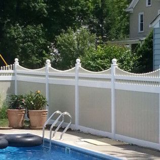 A white and tan fence surrounds a backyard pool, trees and a house in the background.