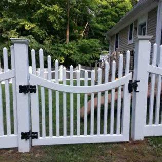 White picket fence with a gate, set in a yard with a house and trees in the background.