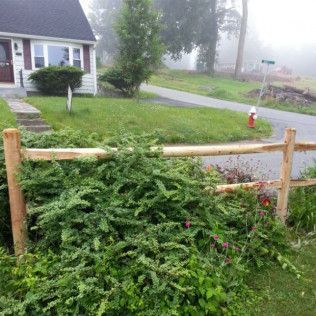 A split-rail fence with lush green bushes in front of a house on a foggy day.