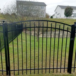 Black metal arched gate in a green yard, with a fence and houses in the background.