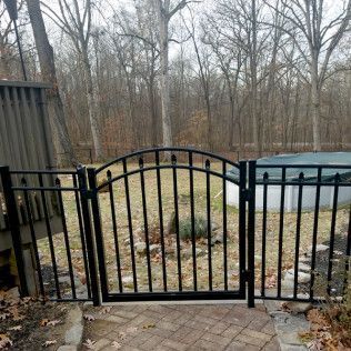 Black metal gate on a brick path leading to a backyard with trees and a pool.