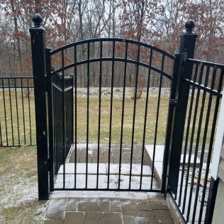 Black metal gate in a yard with a curved top, leading to steps. Snow on the ground, trees in the background.