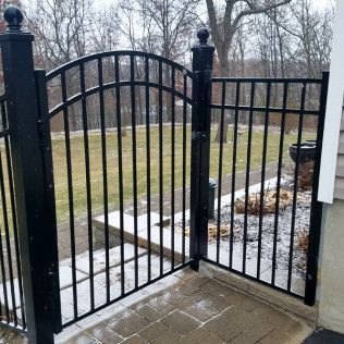 Black wrought-iron gate and fence in front of a lawn and trees. The gate is arched at the top and closed.
