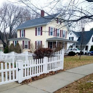 Yellow house with white picket fence, red shutters, leafless trees, and a sidewalk.