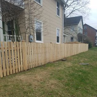 A wooden fence surrounds a beige house on a grassy lawn. Another house is in the background.