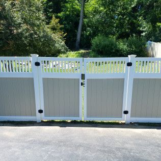 White and beige vinyl fence with a double gate. Black hardware. Trees in background.