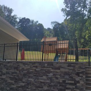 Wooden playhouse behind a black metal fence on a stone wall, backyard setting.