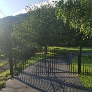 Black gate and fence with a driveway leading toward a large tree and mountain in the background.