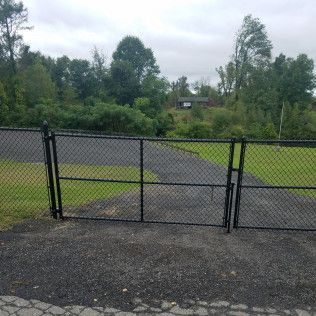 Black chain-link fence with open gate on a gravel driveway, leading to a wooded area under an overcast sky.