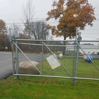Chain-link fence with diagonal supports and a sign hanging, on grass next to a road. A tree is in the background.