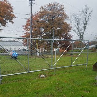 A chain-link gate is open in a green yard. Behind it are trees and buildings under a cloudy sky.