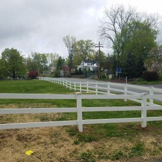 White fence along a grassy field bordering a road with houses and trees in the background on a cloudy day.