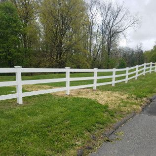 White three-rail fence along green grass, next to a road, with trees in the background.