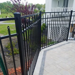 Black metal fence surrounds a gray stone patio, near a white house with green grass.