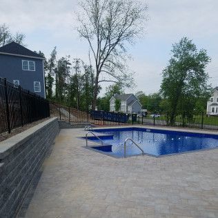 Pool with steps, surrounded by pavers, a black fence, and a retaining wall in a residential area.
