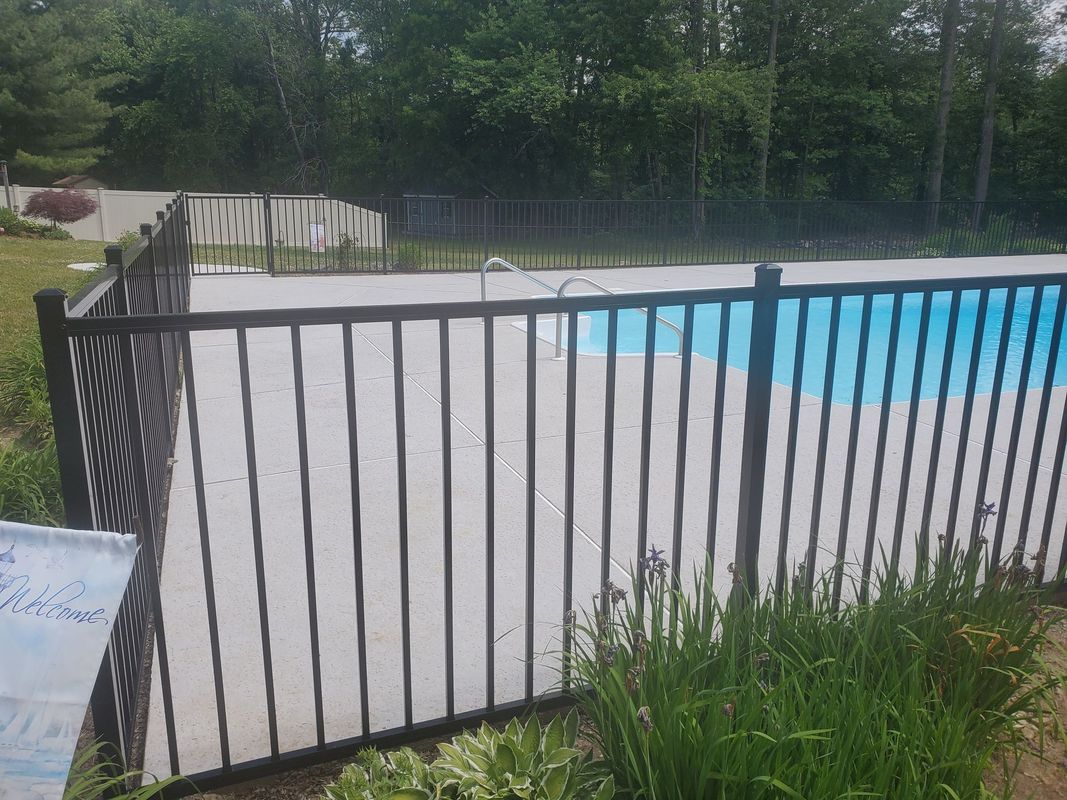 A black fence surrounds a swimming pool covered in a light gray tarp. Green grass and trees.