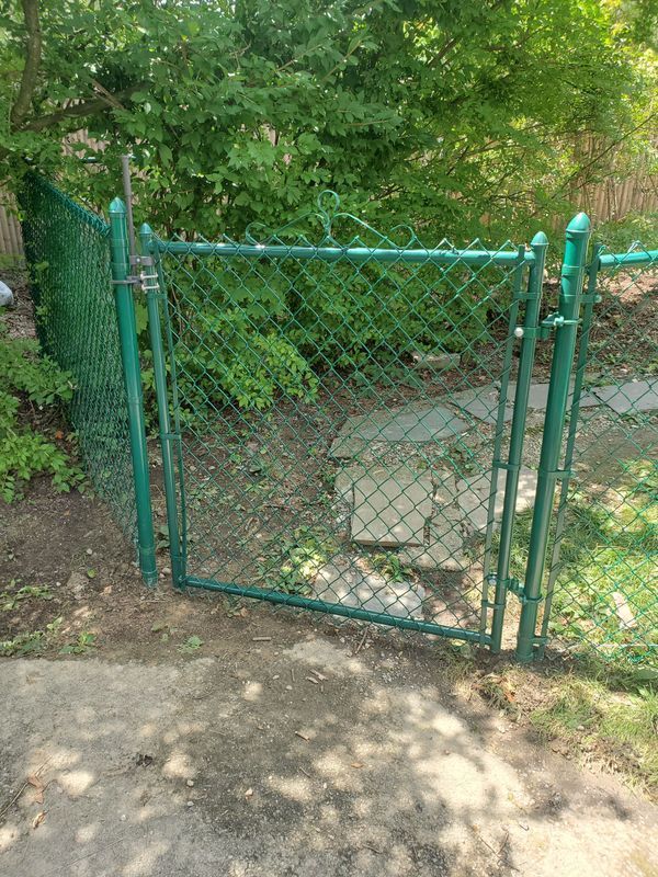 Green chain-link fence with a gate open, leading into a garden area with foliage.