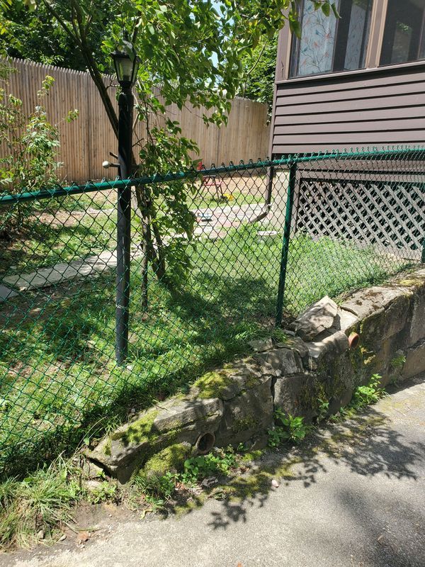 Green chain-link fence bordering a lawn with a stone wall, tree, and house.