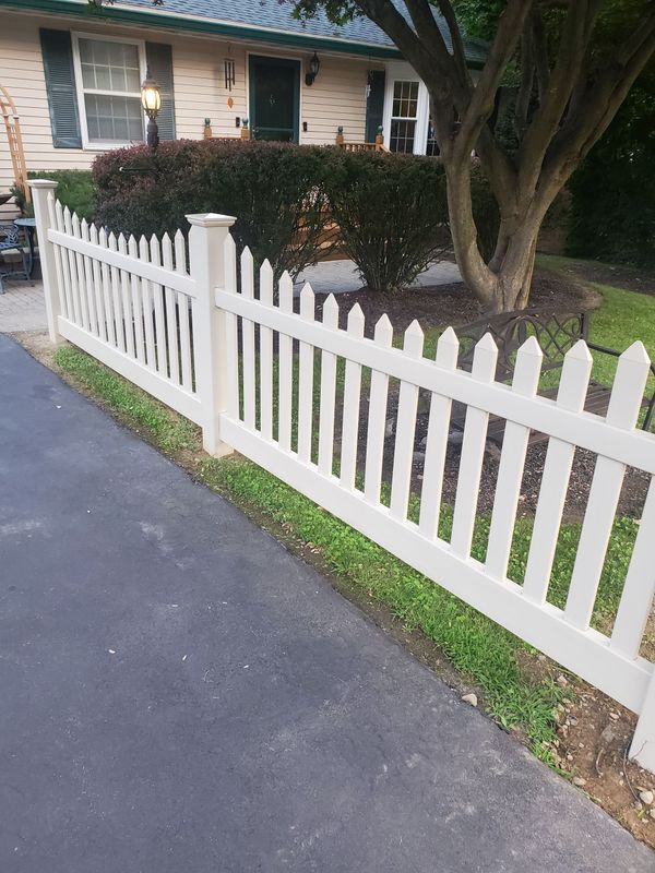 White picket fence bordering a driveway, in front of a house with a small bush and tree.