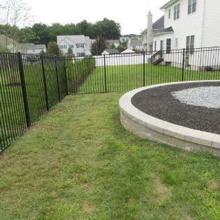 Black fenced backyard with a circular fire pit, green grass, and houses in the background.