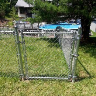 Chain-link fence gate open in a grassy yard, with a pool visible in the background.