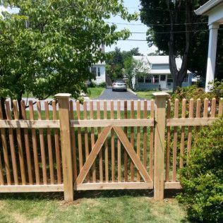 Wooden picket fence with gate in front yard, houses and car in background.