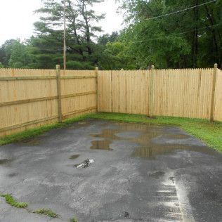 A wet asphalt area surrounded by a wooden fence with grass, trees, and cloudy sky in background.