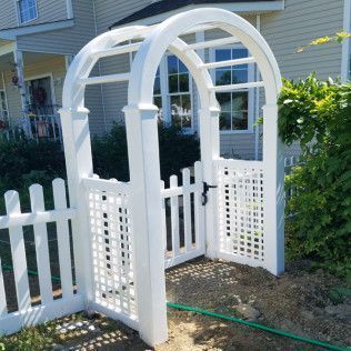 White arched arbor with gate, leading into a garden, with a white picket fence.