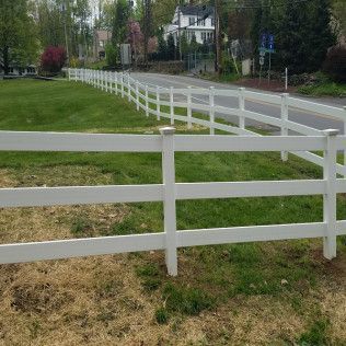 White three-rail fence alongside a grassy roadside, leading toward houses.