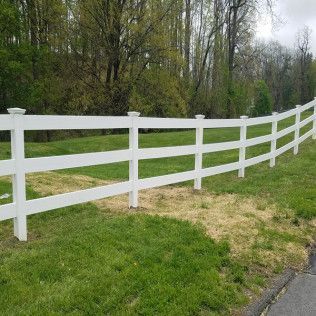 White three-rail fence in a grassy yard, with trees in the background.