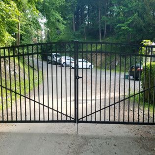 Black metal gate closing a driveway with cars, trees, and road in the background.