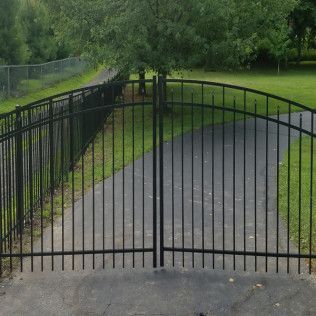 Black metal gates on a paved driveway, with a grassy lawn and trees in the background.