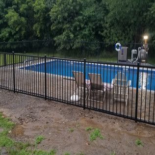Black fenced pool with blue water, lounge chairs, and a wooded background.