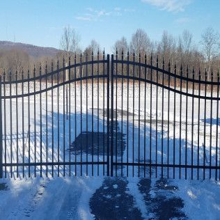 Black metal gate in a snowy field, arched design with a mountain and trees in the background.