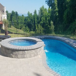 Pool with hot tub, water cascading from tub to the pool, surrounded by patio, fence, and trees.