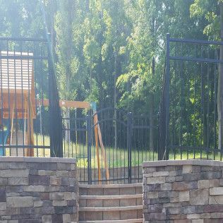Stone wall with steps leading to a black metal fence and gate. In the background are trees and a playset.