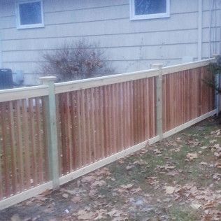 A wooden fence with vertical slats stands in front of a house with windows, on a grassy lawn.